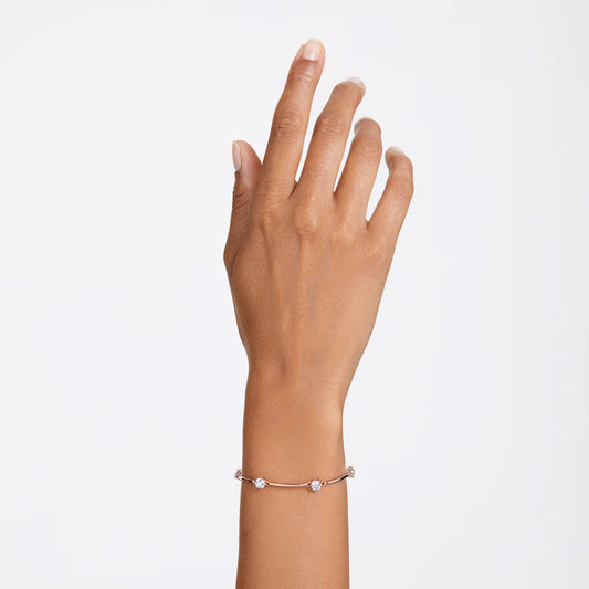 A hand wearing the Swarovski Constella Bangle in a rose gold-tone, featuring small round gemstones and sparkling zirconia, is raised against a plain white background. The bracelet's delicate metal links elegantly connect each stone.