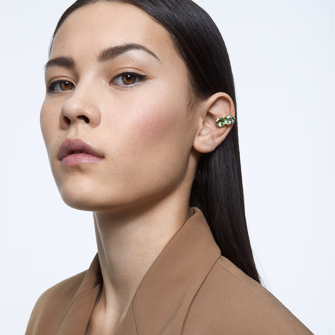 A person with long dark hair and a neutral expression is wearing a brown blazer, accentuated by the Swarovski Millenia Clip Earrings, which are beautifully adorned with shimmering green crystals. The plain white background highlights the subtle elegance of their style.