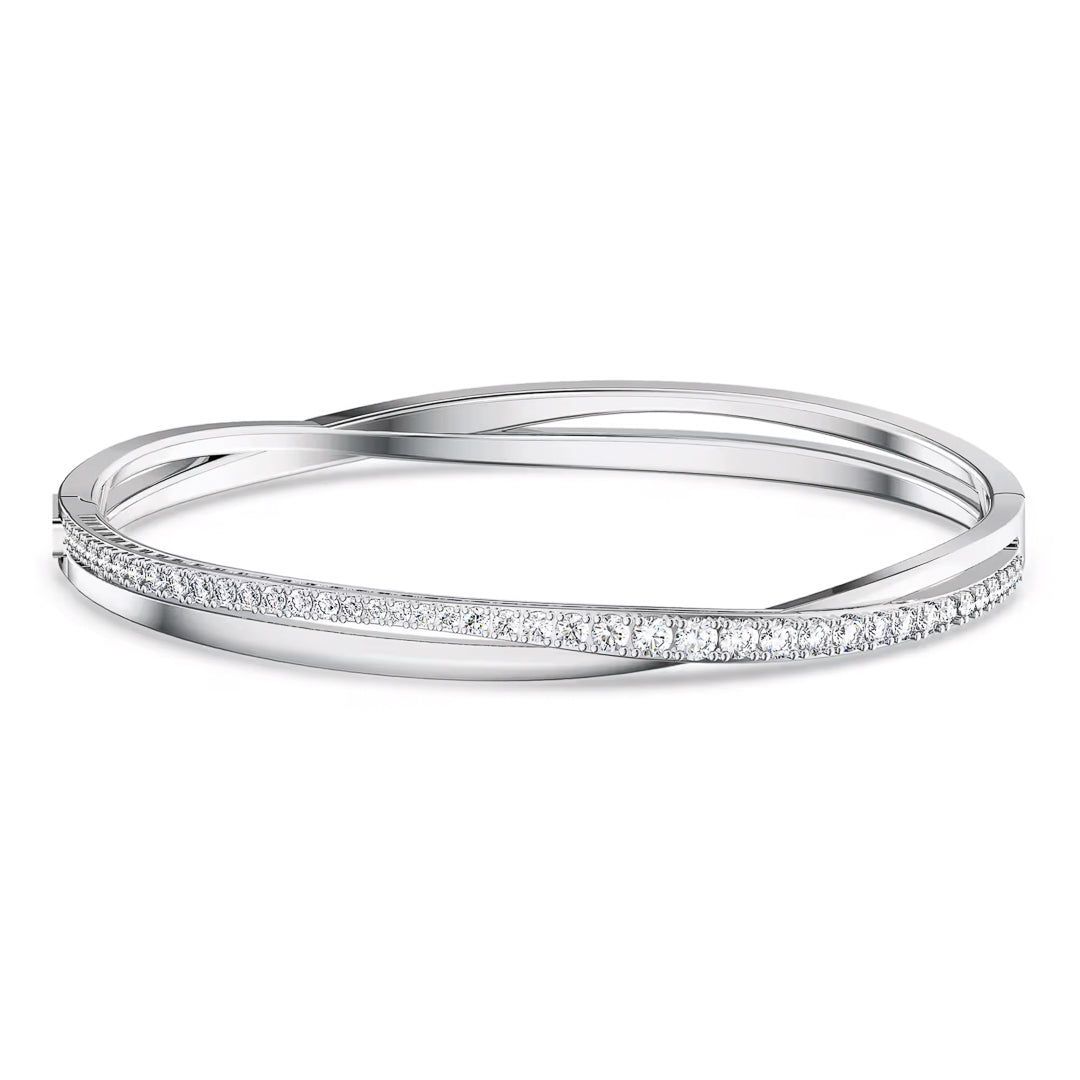The Swarovski Hyperbola Bangle by Swarovski features a sleek silver design with a smooth, polished finish and a row of small, sparkling Swarovski stones embedded along the front. Its elegant and curved design intertwines at one point.