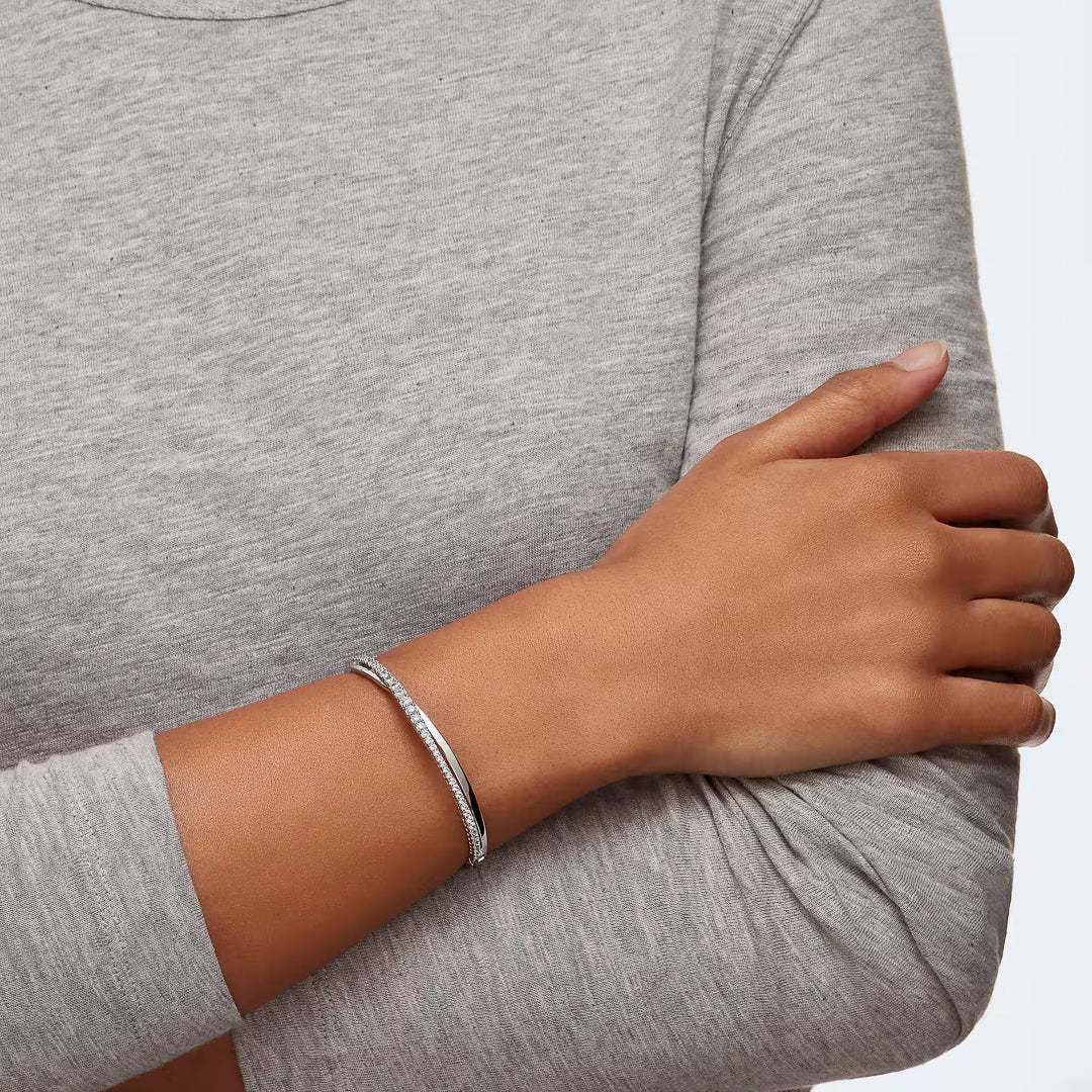 A person in a light gray long-sleeve shirt crosses their arms, showcasing the Swarovski Hyperbola Bangle adorned with zirconia on their right wrist. The plain, light-colored background complements the subtle sparkle perfectly.