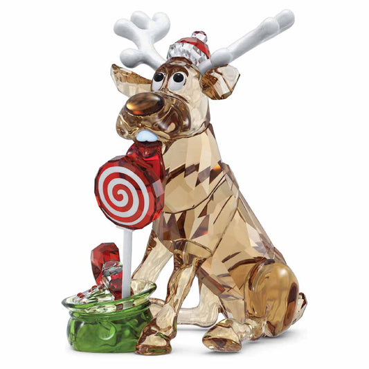 The Swarovski Holiday Cheers Dulcis Reindeer Home Décor from Swarovski is a festive collectible showcasing a whimsical crystal reindeer figurine. It is adorned with antlers, a red hat, and carries a red and white swirl lollipop. Accompanying it is a small green gift bag decorated with a red ribbon.