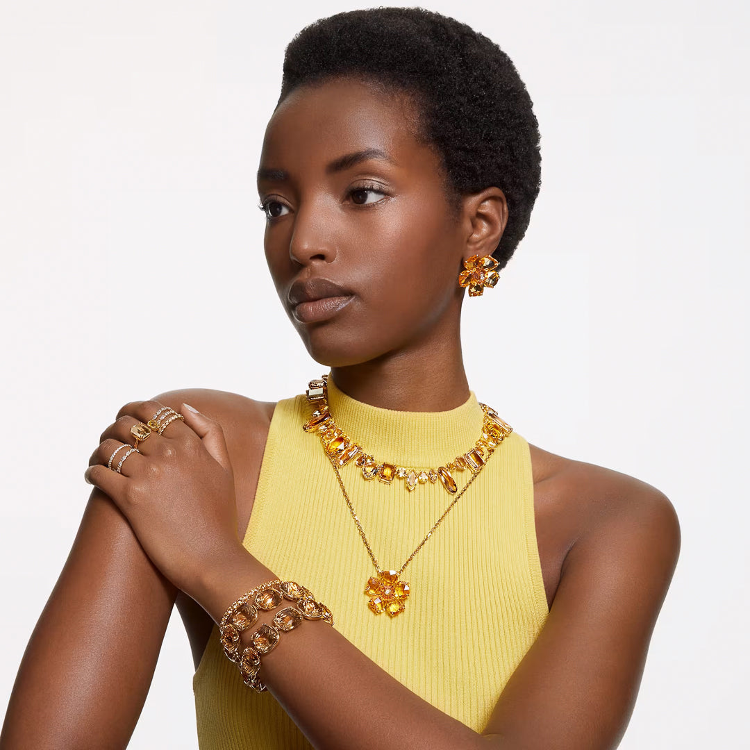 A person wears a yellow sleeveless top complemented by matching jewelry, featuring a Swarovski Vittore Ring. The ensemble includes a chunky necklace, bracelet, earrings, and additional rings, all showcasing a gold-tone finish with striking large orange-gem accents against a simple white background.
