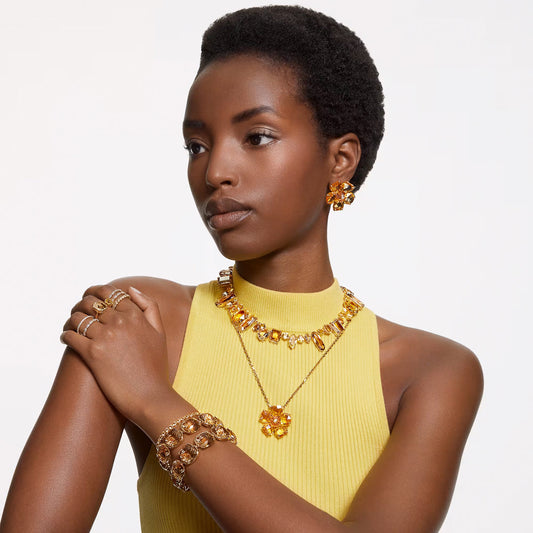 A person wears a yellow sleeveless top complemented by matching jewelry, featuring a Swarovski Vittore Ring. The ensemble includes a chunky necklace, bracelet, earrings, and additional rings, all showcasing a gold-tone finish with striking large orange-gem accents against a simple white background.