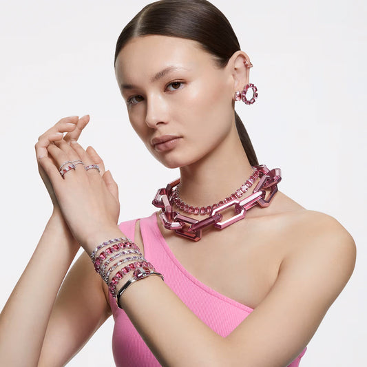 A person with long dark hair wears chunky pink jewelry, including a Swarovski Vittore ring. They are dressed in a one-shoulder pink top adorned with shimmering zirconia accents, posing with hands elegantly crossed against a white background.