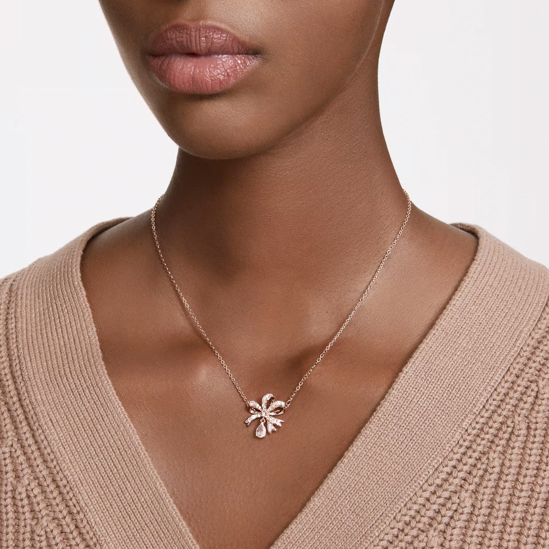 A close-up of a person wearing a beige sweater showcases a delicate Swarovski Hyperbola Necklace Pendant. The rose gold-tone plated piece from the renowned brand, Swarovski, features a pendant shaped like a flower adorned with clear crystals, resting elegantly on the chest. The plain white background highlights the jewelry's brilliance.
