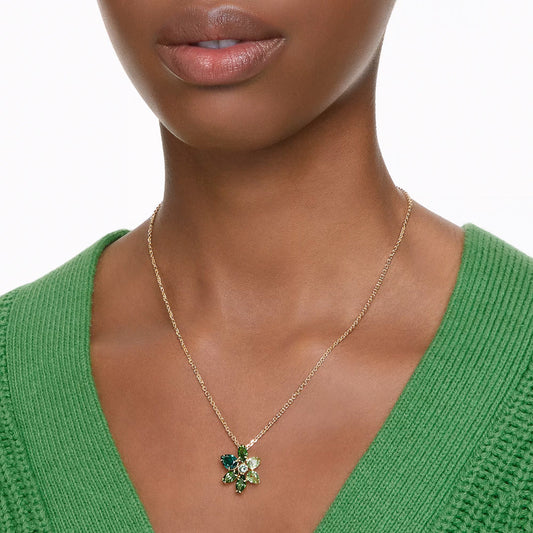 A person is wearing a green textured top with a gold Swarovski Gema Necklace Pendant, which is adorned with facetted crystals and colorful flowers.