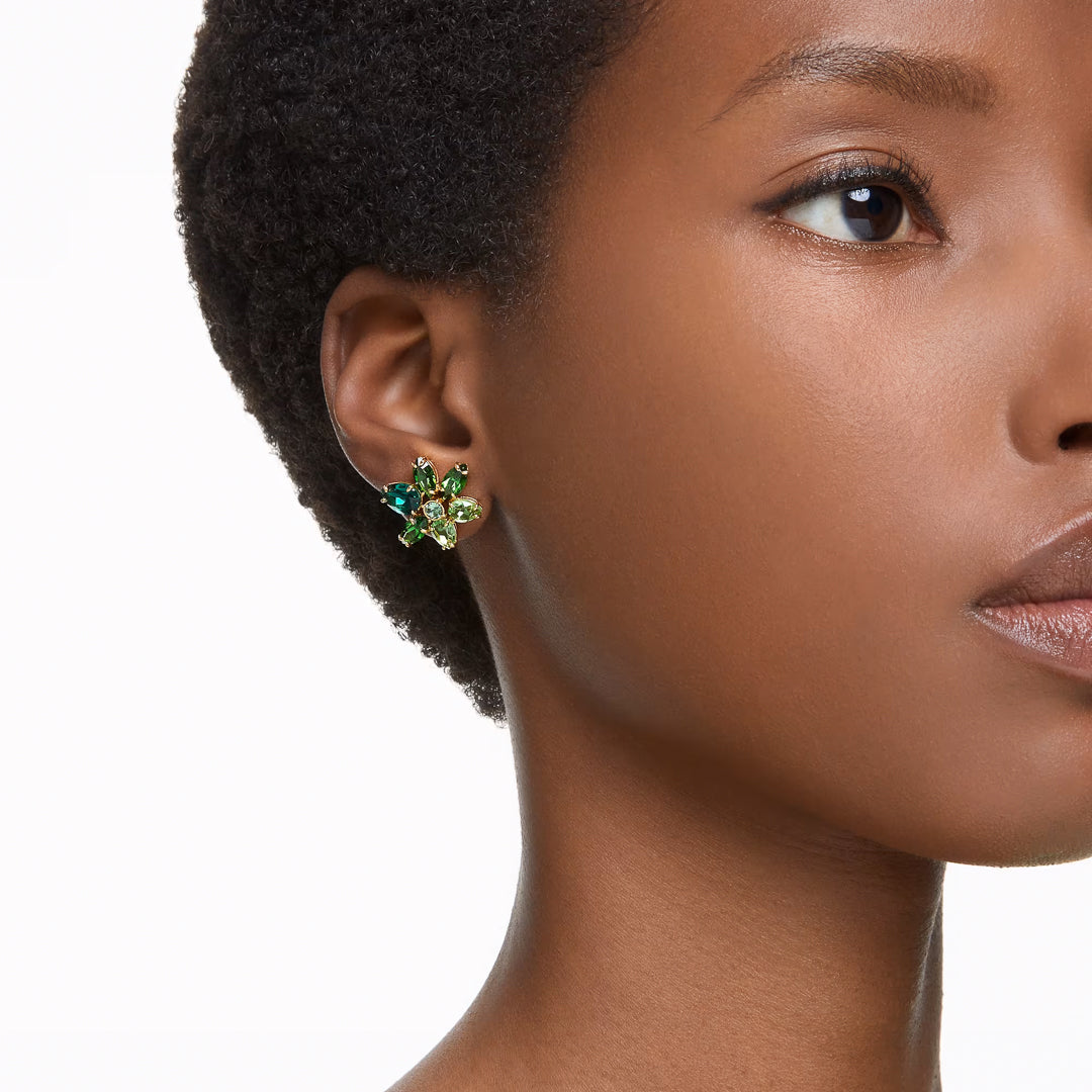 A close-up of a person wearing the sophisticated Swarovski Gema Stud Earrings, featuring green crystals and gold-tone plating. The earrings beautifully contrast with the person's dark skin and short, textured hair, emphasizing a refined and stylish appearance.