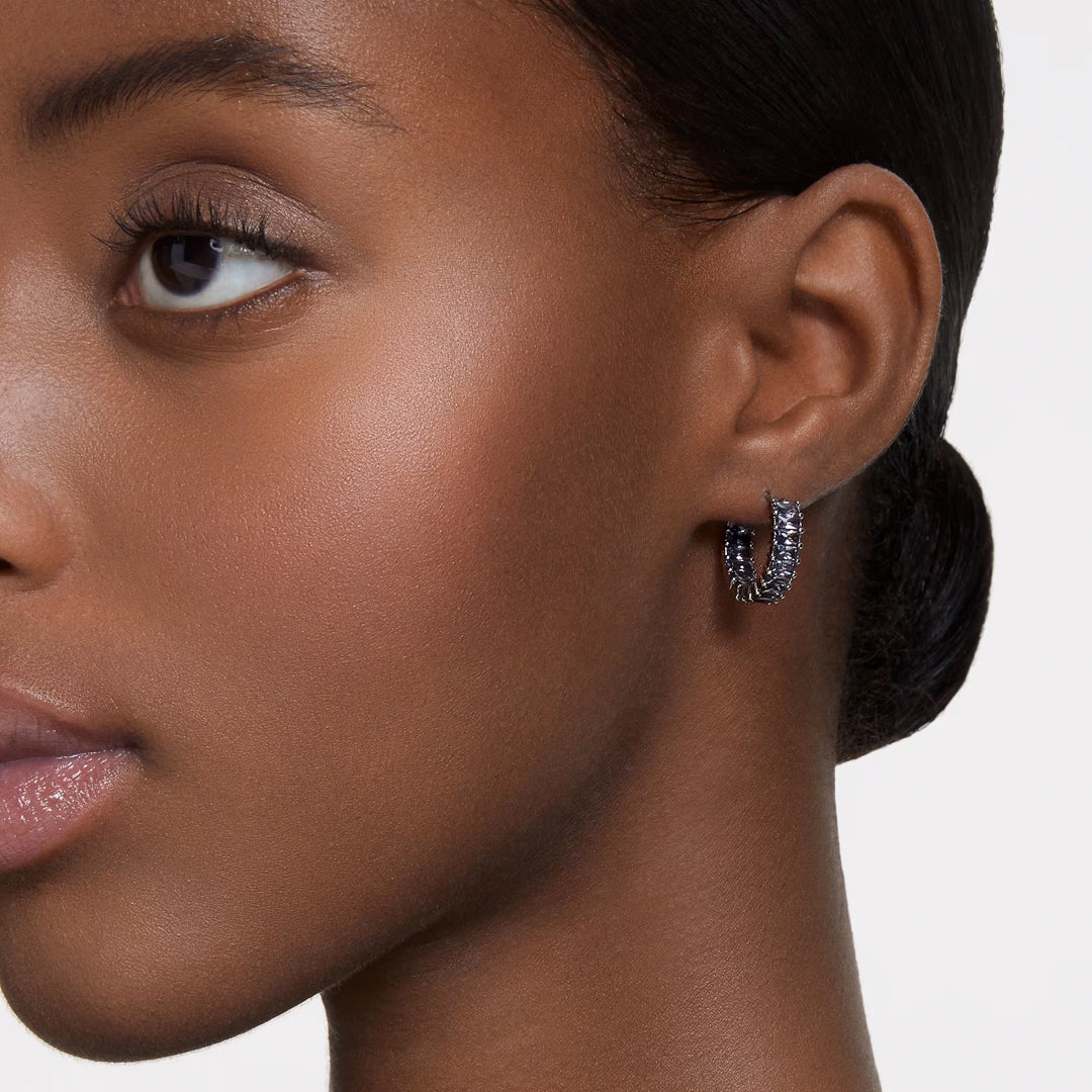 Close-up of the side profile of a person's face, showcasing a single textured Swarovski Matrix Hoop Earring with ruthenium plating. The plain background highlights the person's smooth skin and the striking earring by Swarovski.