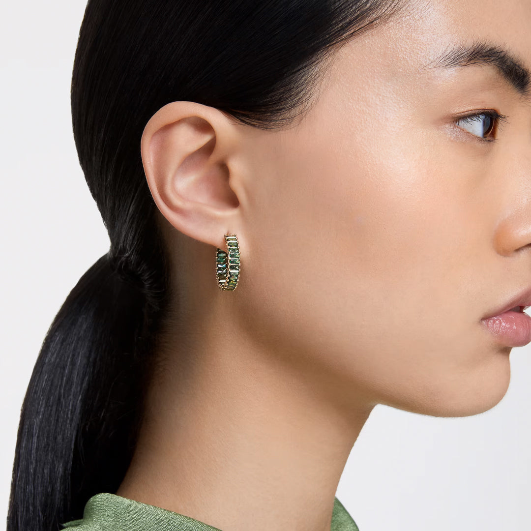 A person with a slicked-back hairstyle is featured in profile, wearing Swarovski Matrix Hoop Earrings adorned with deep green stones. They are also dressed in a green top, and the plain background accentuates the intricate details of the accessory by Swarovski.