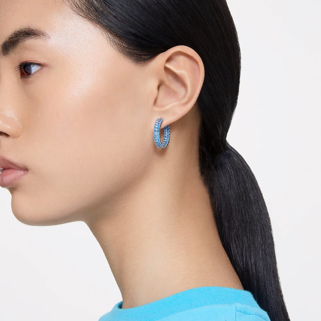 A side profile of a person with long, dark hair tied back showcases them wearing a blue top and a pair of large Swarovski Matrix Hoop Earrings by Swarovski. The plain, light-colored background accentuates the earrings and their sleek hairstyle.