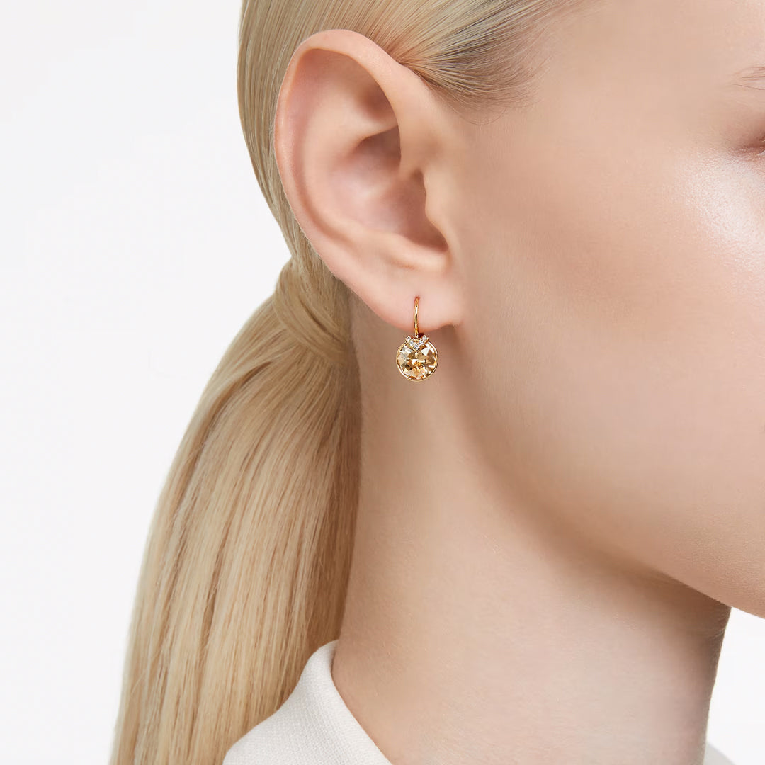 Close-up of a person with long blonde hair tied back, wearing Swarovski Bella V Drop Earrings from Swarovski. These gold-tone earrings feature a round, sparkly pendant with a faceted golden center stone. The person is in a white top and facing sideways, highlighting the clear pavé detailing as well as the earring and hairstyle.