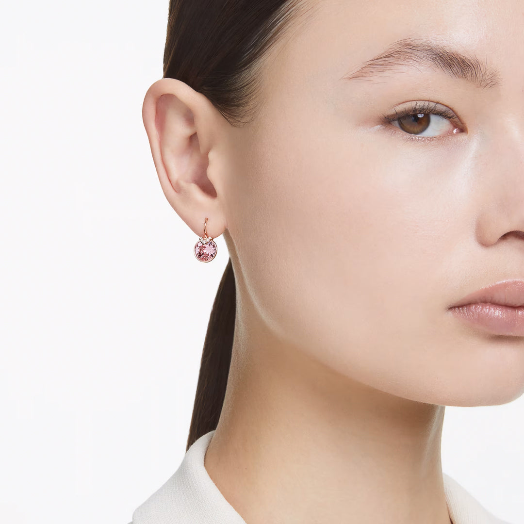 Close-up of a person wearing Swarovski Bella V Drop Earrings, featuring a round, pink stone set in rose gold-tone metal. The plain white background highlights the elegance of the earrings and complements the person's neutral expression.