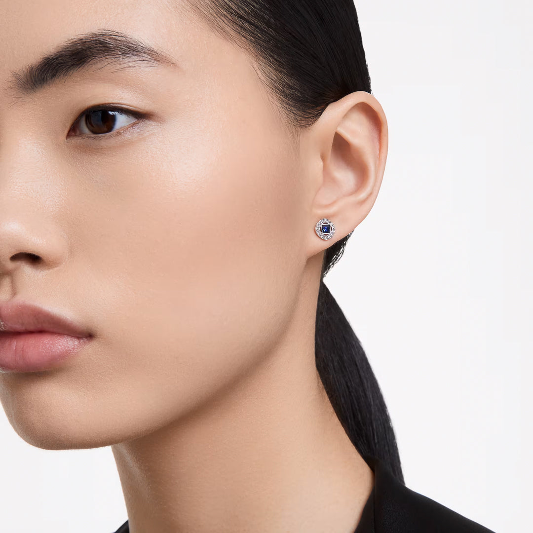 The image focuses on a person with smooth skin and dark hair pulled back, highlighting a Swarovski Angelic Stud Earring with a small, round blue center stone in their left ear. The rhodium-plated design from Swarovski adds elegance against the simple, light-colored background.