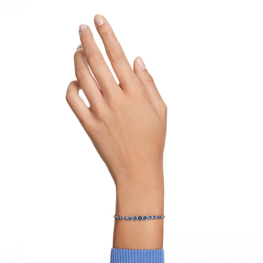 A hand adorned with the Swarovski Imber Emily Bracelet, a rhodium-plated silver piece featuring shimmering blue crystals. The wearer, with a light skin tone, is elegantly dressed in a blue long-sleeve top against a plain white backdrop.