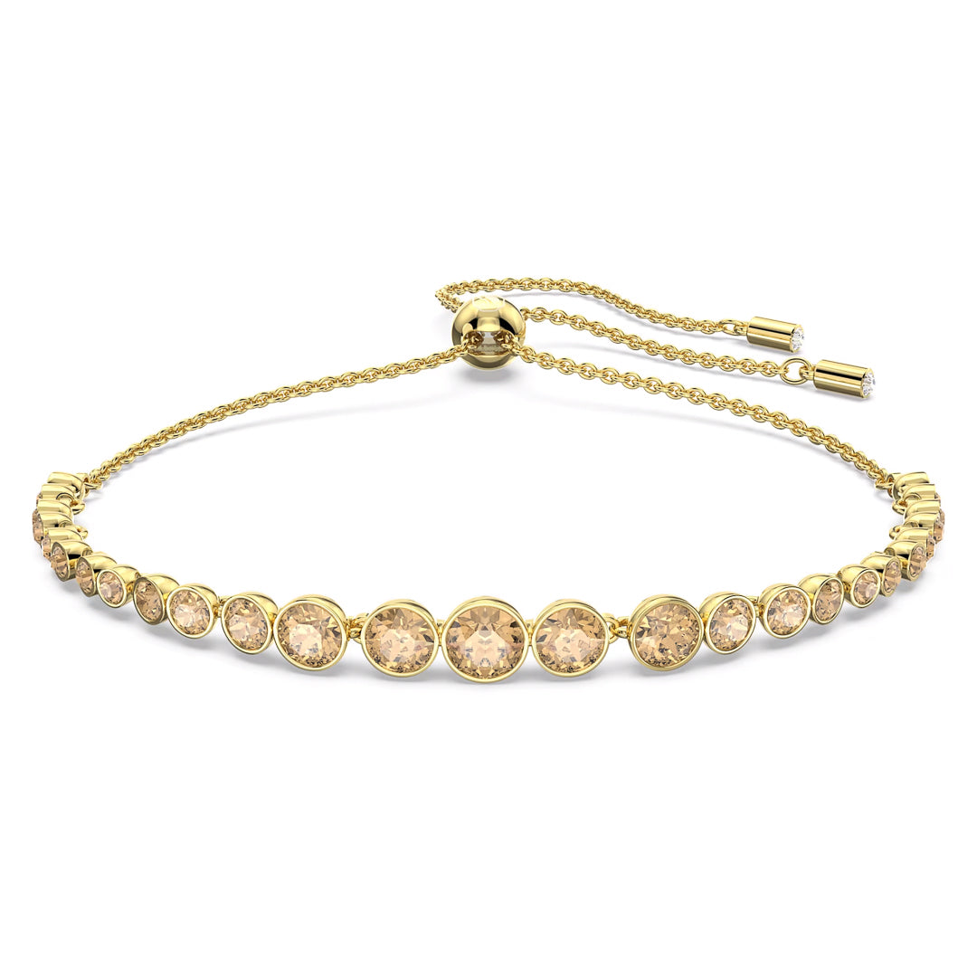The Swarovski Imber Emily Bracelet is a refined gold-tone accessory showcasing graduated golden crystals arranged in a row. This sophisticated piece is equipped with a sliding clasp and an adjustable chain featuring two dangling ends, each embellished with a small gemstone.