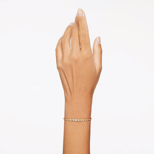 A hand wearing the elegant Swarovski Imber Emily Bracelet, featuring a delicate gold-tone with evenly spaced small white stones, is held up against a plain white background. The sliding clasp adds functionality, while the bracelet's simplicity and elegance are beautifully highlighted by the uncluttered setting.