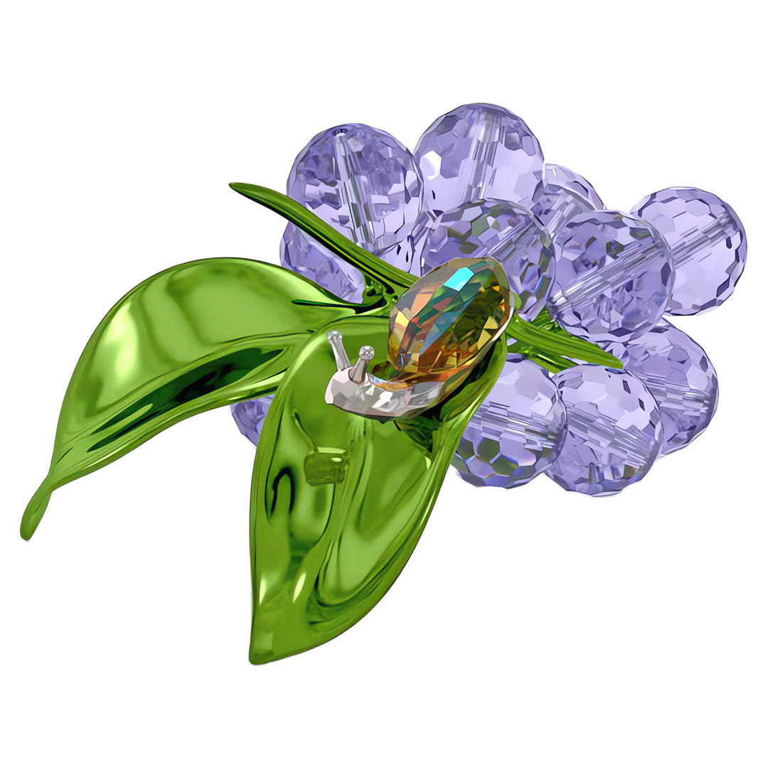 The Swarovski Idyllia Snail and Blueberries Home Décor showcases a cluster of transparent purple grapes crafted from faceted crystals, adorned with bright green, shiny leaves. It includes a small, detailed crystal snail perched on the grapes, featuring a clear shell and a golden body.