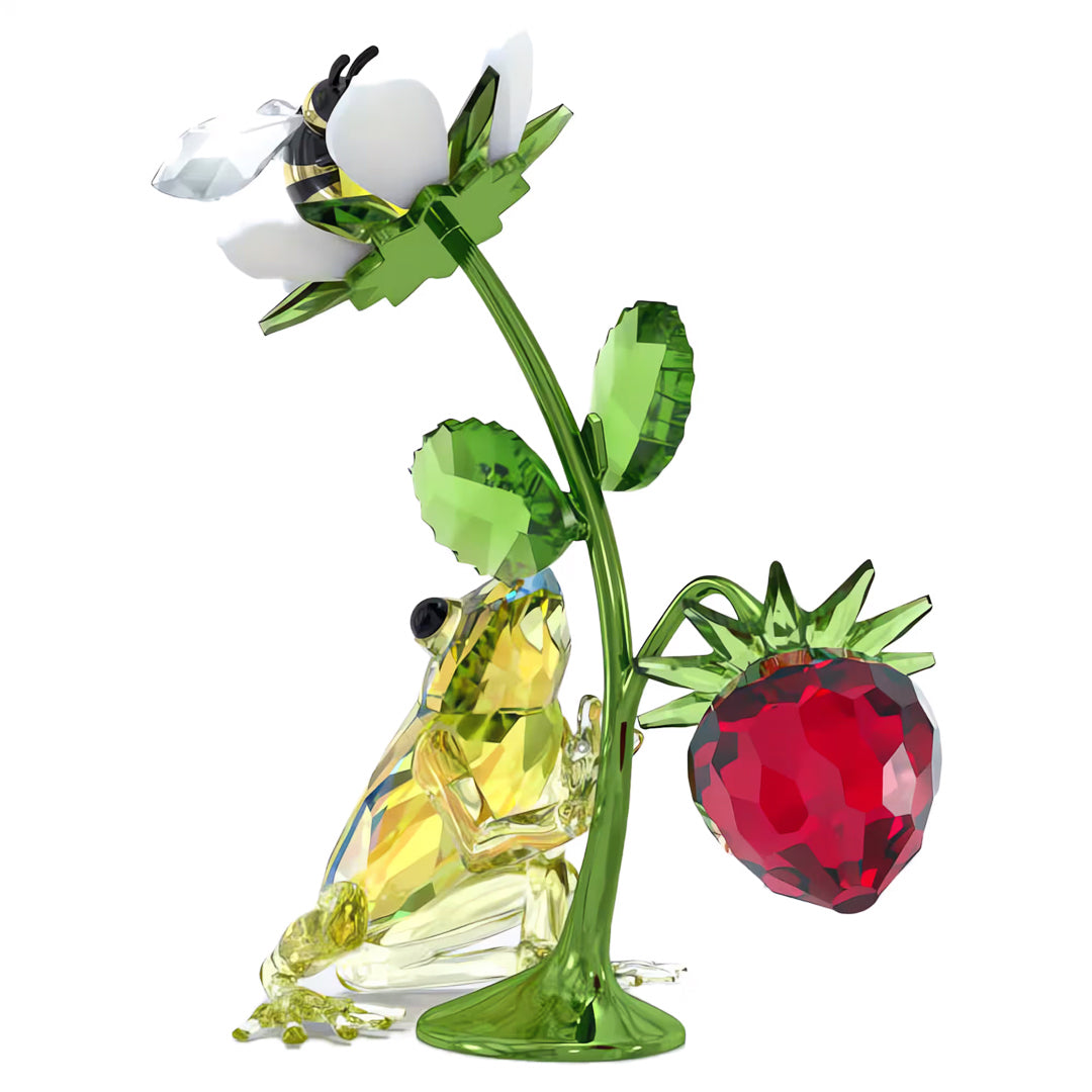 The Swarovski Idyllia Frog Bee and Strawberry Home Décor showcases a crystal frog resting against a green stem, accompanied by the vibrant red fruit of a strawberry branch. Above, a white flower adorned with a bee sits gracefully, all intricately crafted in Swarovski's signature faceted design.