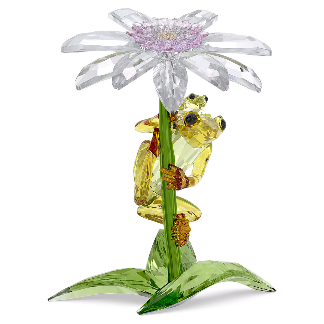 The Swarovski Idyllia Frogs and Flower Home Décor piece showcases a crystal sculpture of a yellow frog clinging to a green stem, topped with a large, translucent flower adorned with purple and clear Swarovski crystal petals. The base is crafted from green leaves, while the frog is accentuated by shiny brown feet. This stunning piece instantly adds an air of tranquillity to any home décor setting.