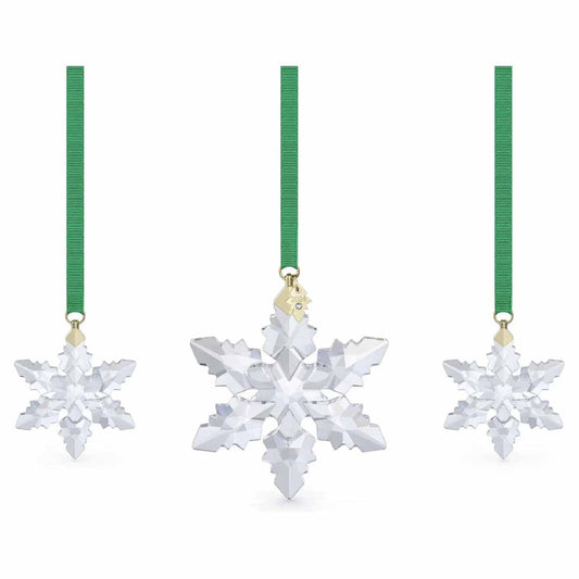 Three Swarovski 2024 Annual Edition Ornaments are suspended by green ribbons against a white background. Each ornament embodies intricate facets and clear, reflective surfaces, characteristic of crystal snowflakes.