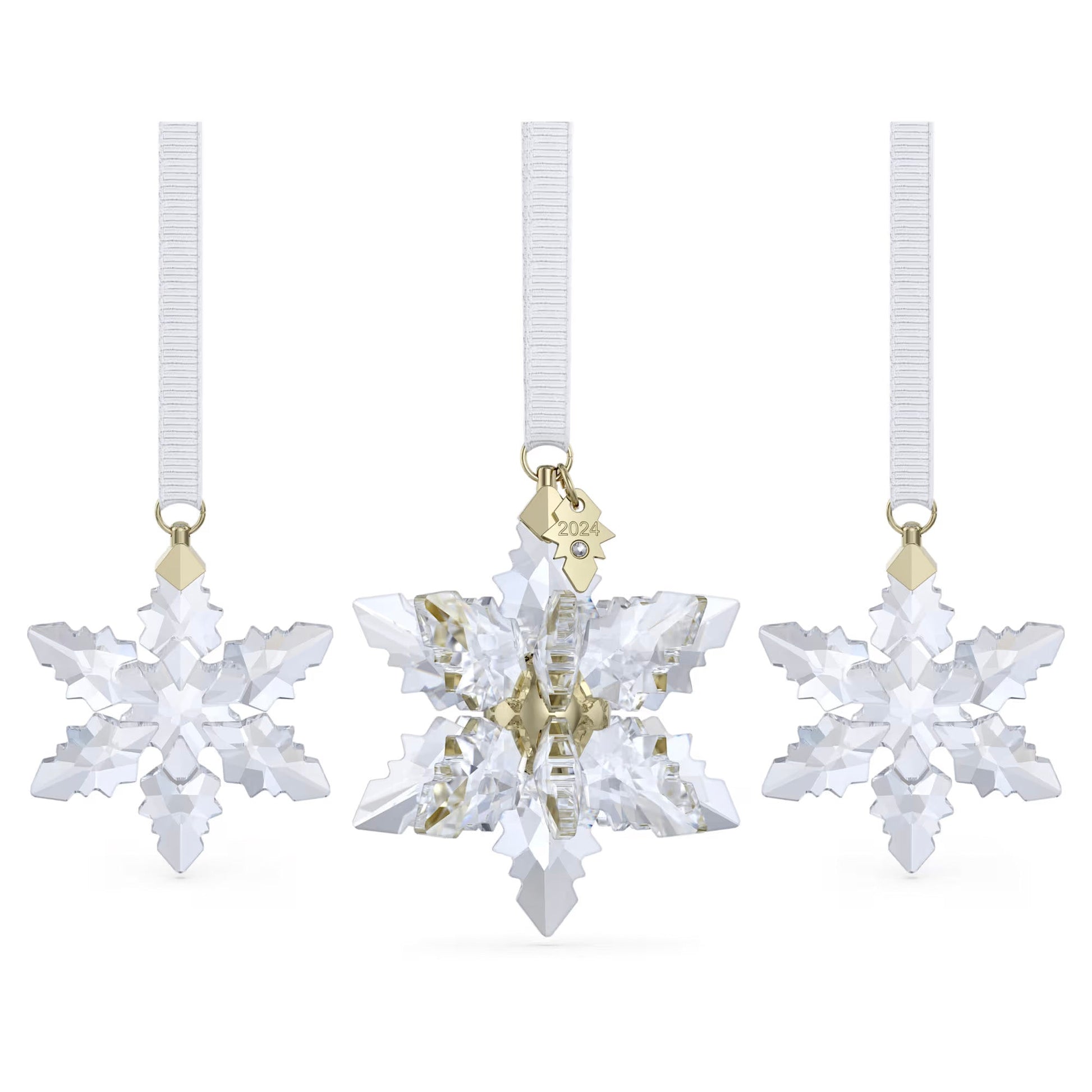 Three crystal snowflake ornaments hang from white ribbons, with the center one being a larger piece from the Swarovski Annual Edition 3D Ornament Set 2024. This middle ornament is adorned with a small gold tag inscribed "2024." Each ornament in the set sparkles with intricate facets, reflecting light beautifully and adding to the festive atmosphere.