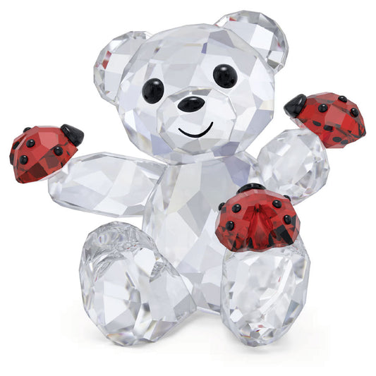 The Swarovski Kris Bear Good Luck Bear Home Décor, crafted by Swarovski, is a joyful figurine made of sparkling crystals. It sits on a white background while holding two charming red crystal ladybugs with black spots on its paws.