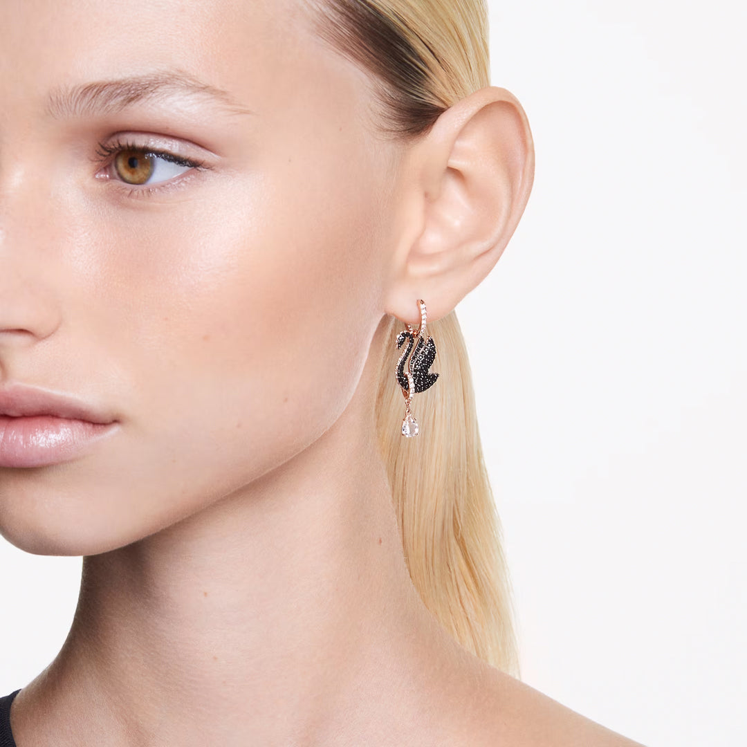 A close-up shot features a person with light-colored hair wearing Swarovski Swan Drop Earrings. The striking black swan motif, enhanced by a dangling white gem and rose gold-tone details, is prominently displayed against the plain white background, highlighting its intricate craftsmanship.