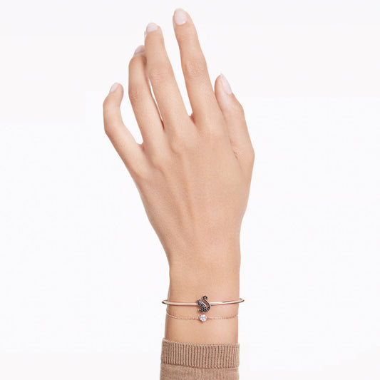 A hand is depicted wearing two delicate bracelets on a light skin tone. One bracelet, the Swarovski Swan Bangle by Swarovski, showcases a dazzling swan motif with multicolored accents in a rose gold-tone. The second bracelet is a simple chain featuring a small, round charm. The individual is dressed in a beige long-sleeved top.
