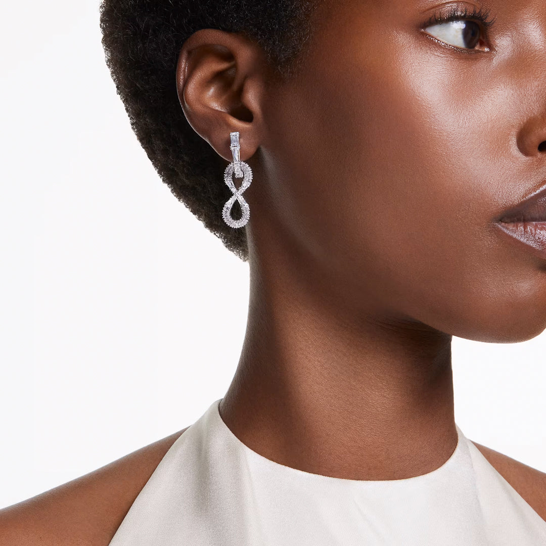 Close-up of a person wearing elegant Swarovski Hyperbola Drop Earrings from Swarovski, shaped like infinity symbols and adorned with sparkling zirconia. The person has short, curly hair and is wearing a white halter neck top. The background is plain white.