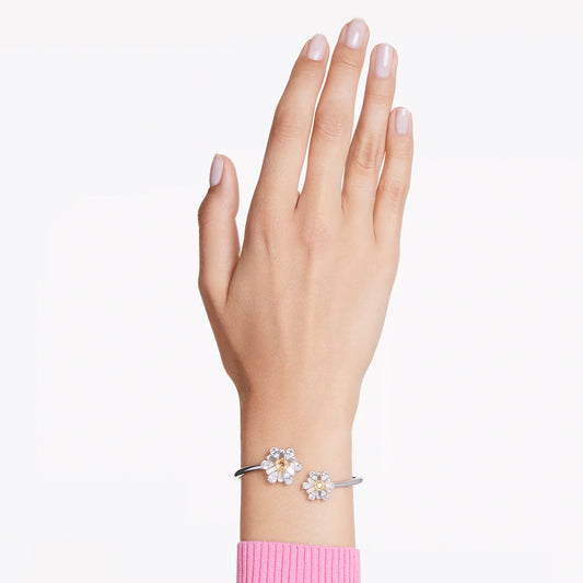 A woman's hand with light pink nails showcases a Swarovski Idyllia Bangle, rhodium-plated and embellished with two flower-shaped ornaments boasting white petals and gold centers. She is dressed in a pink sleeve against a plain white background.
