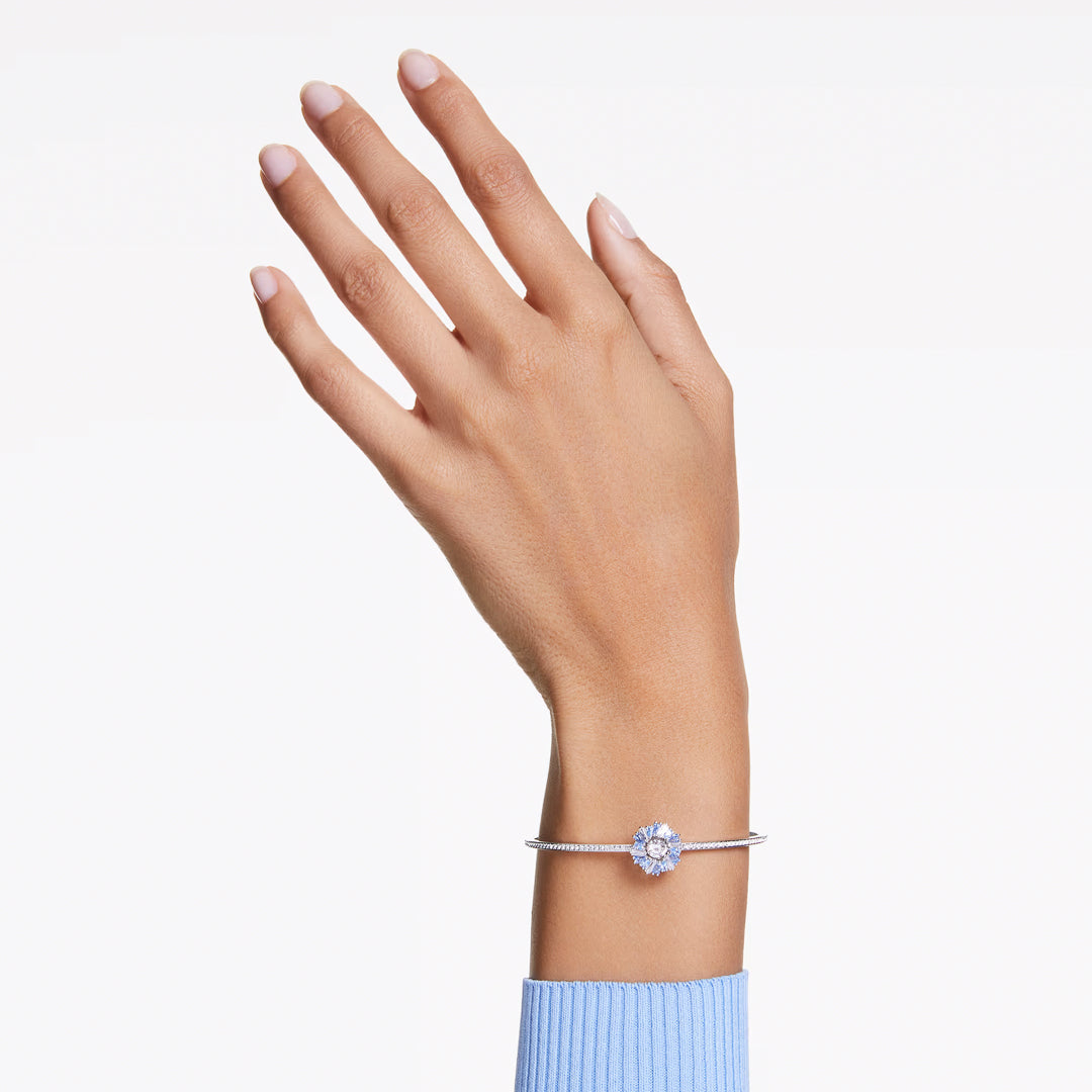 A hand in a blue sweater sleeve appears against a white backdrop, adorned at the wrist with the Swarovski Idyllia Bangle. The bangle features a central circular Swarovski Zirconia gemstone, while the fingers are slightly spread to display a tidy manicure.