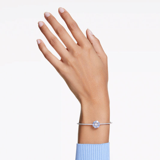 A hand in a blue sweater sleeve appears against a white backdrop, adorned at the wrist with the Swarovski Idyllia Bangle. The bangle features a central circular Swarovski Zirconia gemstone, while the fingers are slightly spread to display a tidy manicure.