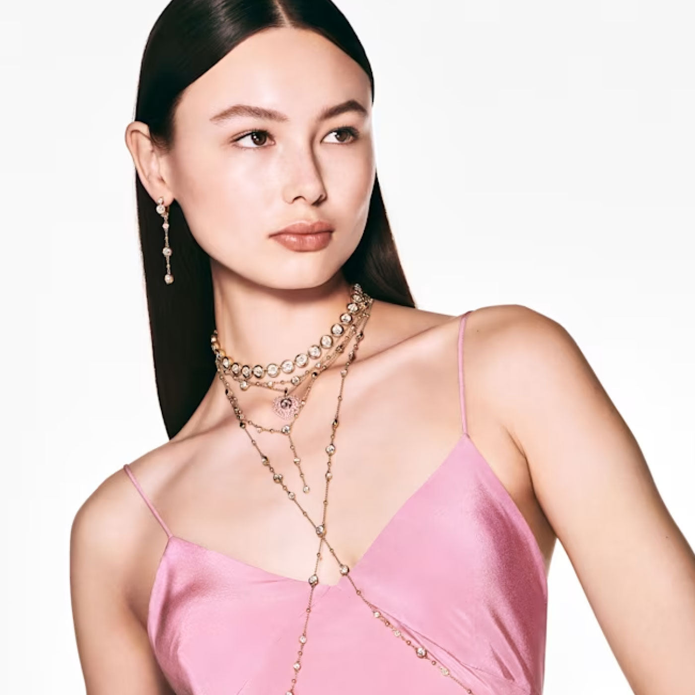 A woman with long dark hair wears a pink satin dress and layered necklaces, gazing to the side. The minimal background emphasizes her elegant jewelry, showcasing the Swarovski Imber Station Necklace, which adds a touch of sophistication to her gold-tone plated attire.