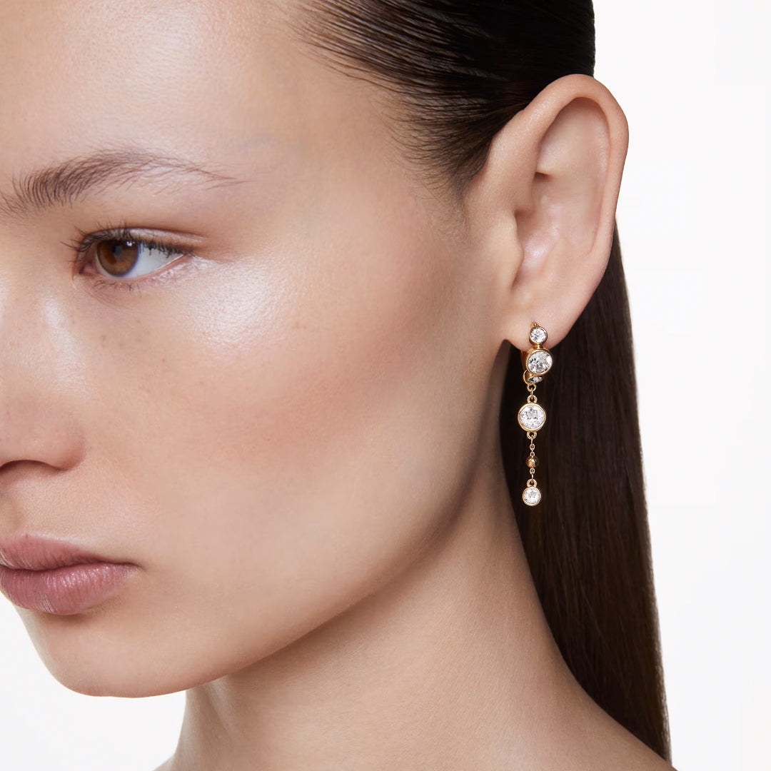 A close-up of a woman wearing Swarovski Imber Drop Earrings shows the elegant gold-tone plated earrings, featuring a delicate chain adorned with three round-cut crystals. Her slicked-back hair highlights the contrast between the earrings and her smooth, natural skin against a plain white background.