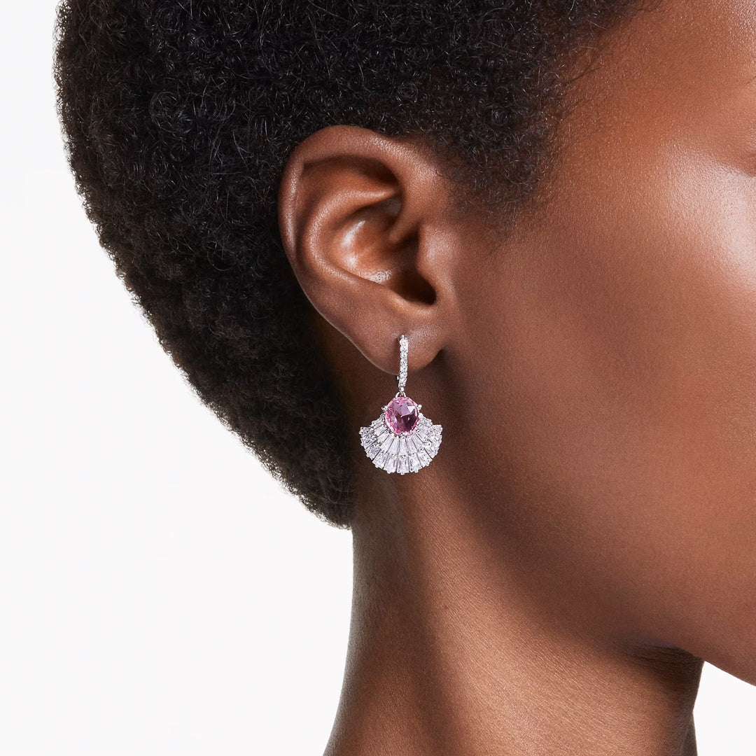 Close-up of an ear wearing the sophisticated Swarovski Idyllia Drop Earring. The piece showcases a pink gemstone embraced by a fan of sparkling white Swarovski Zirconia, creating an elegant contrast against the person's dark skin and short, curly hair.