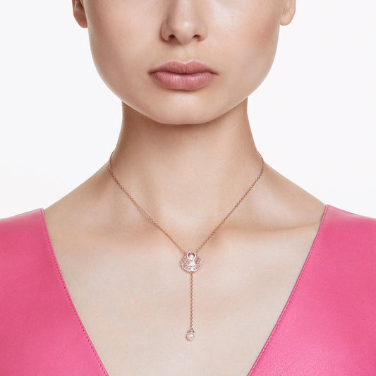 A woman in a pink V-neck top is adorned with the Swarovski Idyllia Y Necklace Pendant, a delicate piece plated in rose gold-tone. The necklace features an ocean-inspired pendant and shimmering Swarovski Zirconia, adding an elegant touch to her ensemble against a plain white background.