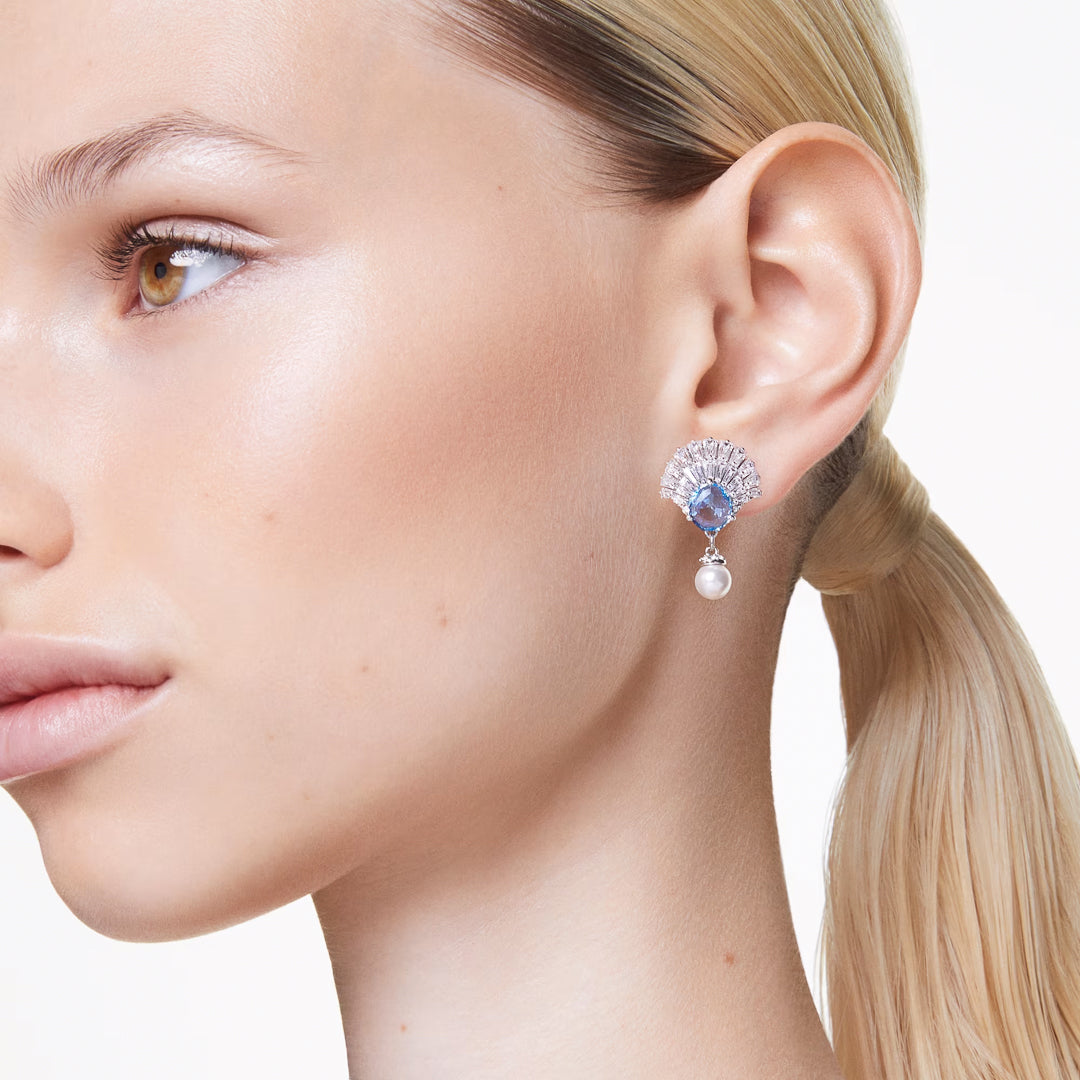 A close-up shot focuses on a person with blonde hair tied back, highlighting the exquisite Swarovski Idyllia Drop Earrings. These elegant accessories boast a fan-shaped design adorned with blue gemstones and sparkling Swarovski Zirconia, complemented by a dangling pearl. Set against a plain white background, these earrings from Swarovski stand out beautifully.