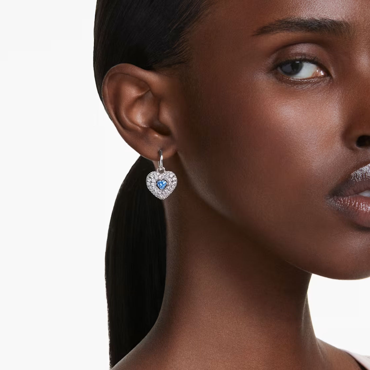 A woman with long, dark hair is wearing Swarovski's Hyperbola Blue Heart Drop Earrings. The background is plain white, emphasizing the shimmering jewelry as only the side of her face is visible.