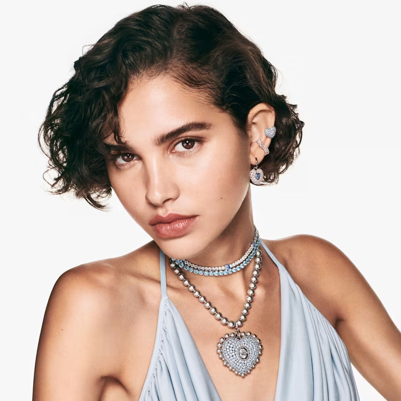 A woman with short, curly hair is wearing elegant jewelry, including Swarovski Hyperbola Blue Heart Drop Earrings, a heart-shaped necklace adorned with Swarovski Zirconia, and a beaded choker. She is dressed in a light blue sleeveless top and looks directly at the camera.