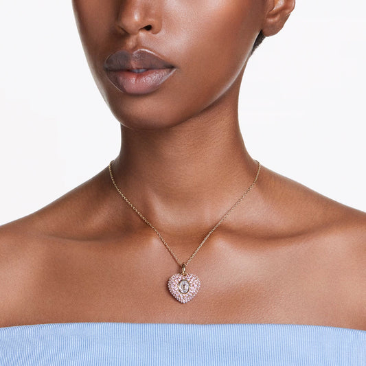 A person wearing a pink Swarovski Idyllia Necklace Pendant with an "O" on a delicate gold chain. The person is wearing a light blue, off-shoulder top against a plain white background.