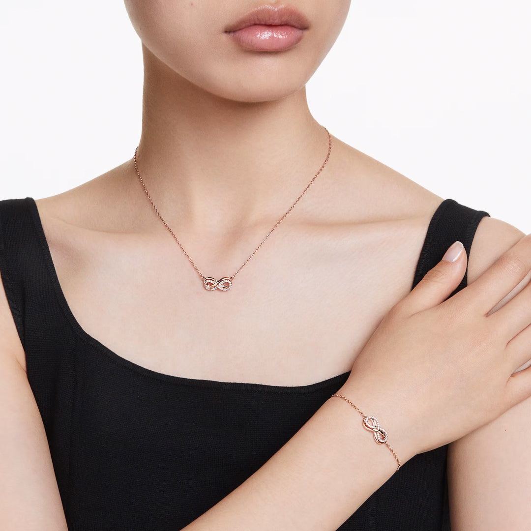 A person wearing a black sleeveless top showcases the Swarovski Hyperbola Necklace Pendant and Bracelet Set in a delicate rose gold-tone, featuring an elegant infinity symbol. The Swarovski jewelry beautifully highlights the timeless infinity theme with its simplicity and elegance.