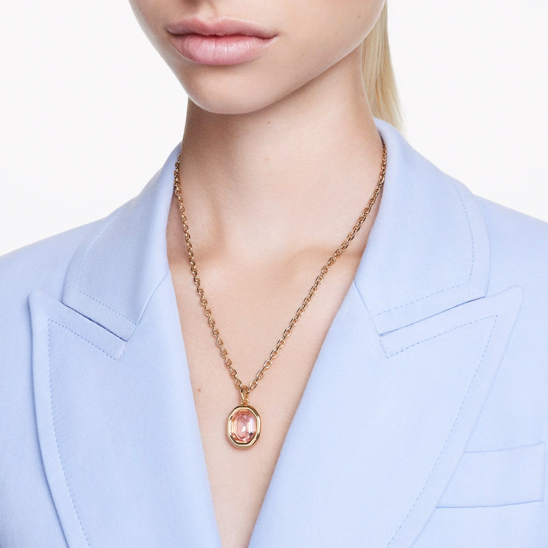 A person wearing a light blue blazer and the Swarovski Imber Necklace Pendant, featuring a gold chain with an octagon-cut pink crystal. The white background accentuates the gold-tone elegance of this Swarovski piece.