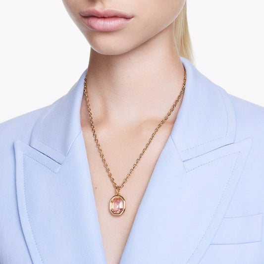 A person wearing a light blue blazer and the Swarovski Imber Necklace Pendant, featuring a gold chain with an octagon-cut pink crystal. The white background accentuates the gold-tone elegance of this Swarovski piece.