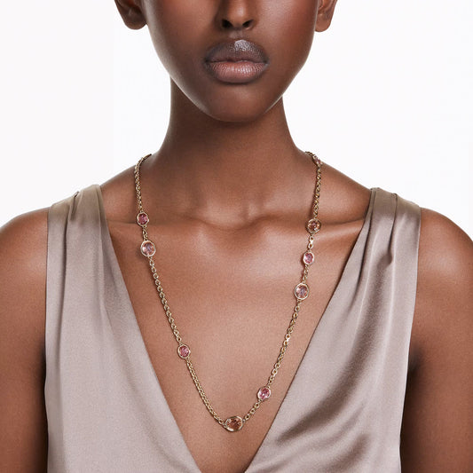 A woman is styled in the Swarovski Imber Strandage Necklace, a long gold chain featuring pink Swarovski Zirconia. She pairs it with a sleeveless, satin-finish taupe top against a plain white backdrop.
