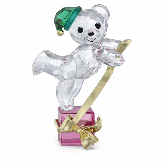 The Swarovski Kris Bear Holiday 2024 Annual Edition Home Décor is a charming collectible that showcases a crystal teddy bear in an endearing pose. It perches on a pink gift box, adorned with a green hat and holding a gold ribbon, while its clear surface beautifully reflects light.