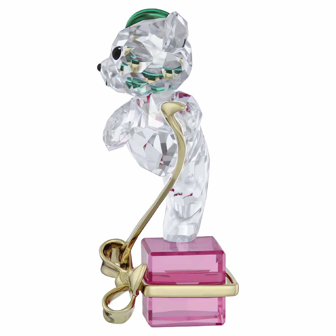 The Swarovski Kris Bear Holiday 2024 Annual Edition Home Décor, a charming crystal figurine adorned with a green cap, is elegantly positioned on a pink base. This collectible features a golden bow wrapped around the base, perfectly blending playfulness with sophistication.