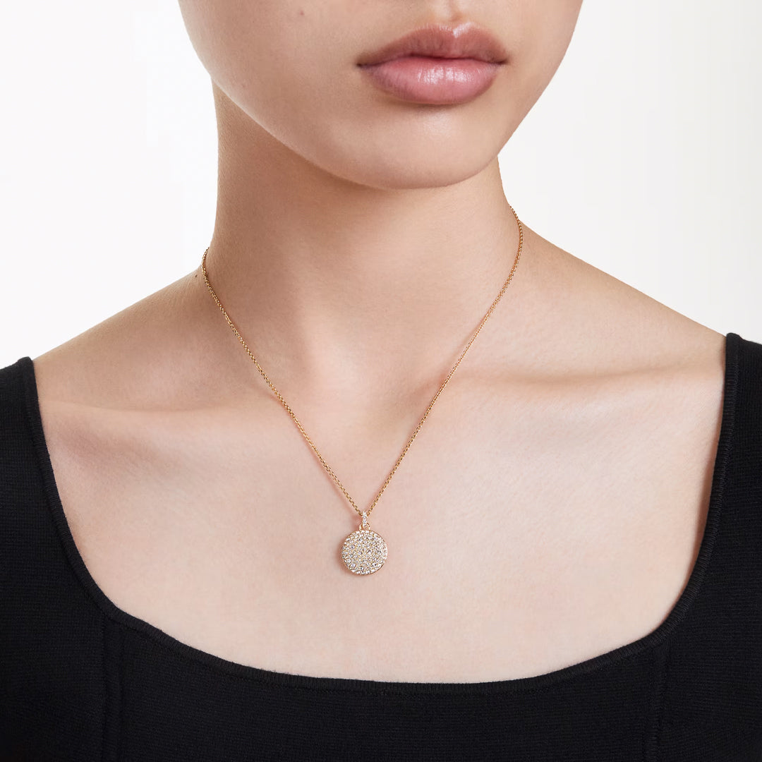A close-up of a person wearing a black square-neck top and the elegant Swarovski Sublima Necklace Pendant, designed with a galaxy-inspired theme and featuring clear round crystals. The softly blurred background enhances the pendant's sparkle, highlighting its exquisite detail from Swarovski.