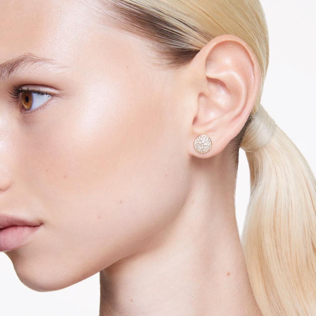 A close-up of a person with light blonde hair pulled back, showcasing the side profile with Swarovski Sublima Stud Earrings. The white background highlights the galaxy-inspired design of these round, pierced earrings from Swarovski.