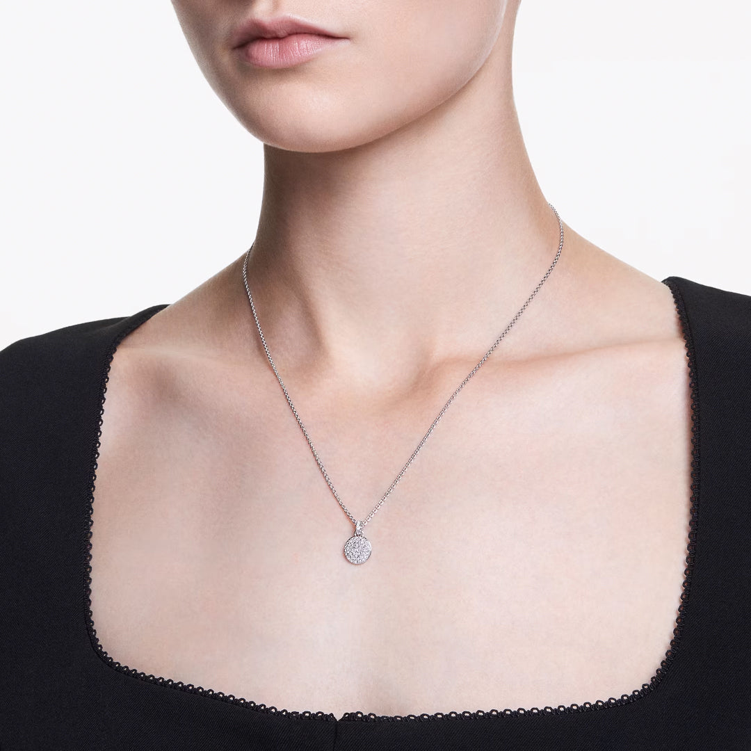 A person wearing a black dress with a square neckline is adorned with the Swarovski Sublima Necklace Pendant, featuring a galaxy-inspired design and clear round crystals. The background is plain white.