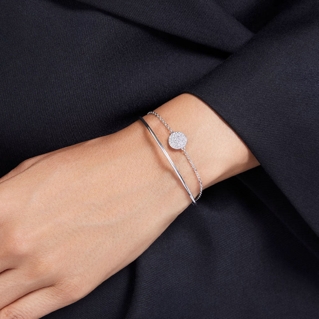 A hand adorned with two silver bracelets: the Swarovski Sublima Bangle, featuring a starry design, and a delicate chain bracelet with a textured circular charm. The rhodium-plated surfaces of both pieces gently catch the light as they rest against the dark fabric background.